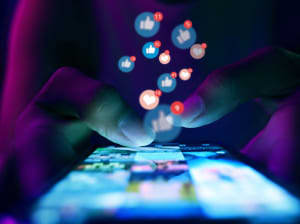 A close up on a smartphone with fingers tapping on a grid of images across the screen. Social media notification icons are floating up from where the screen is being tapped.
