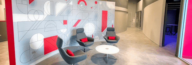 Three gray lounge chairs placed in front of a white wall featuring line-designs. A round coffee table sits in front of the chairs.